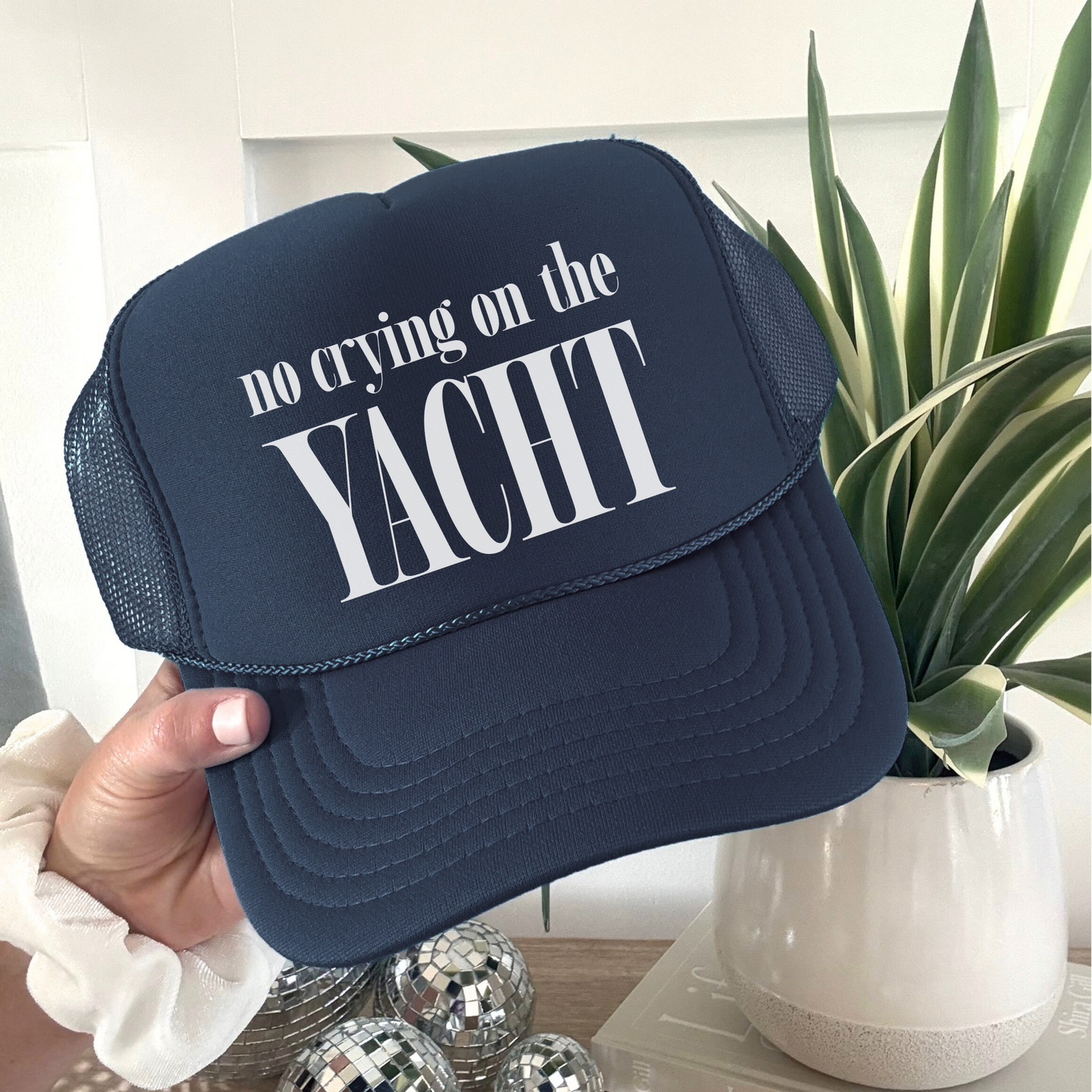 Navy blue trucker hat with white “No Crying on the Yacht” lettering held in hand indoors next to modern plant decor, casual lifestyle photo