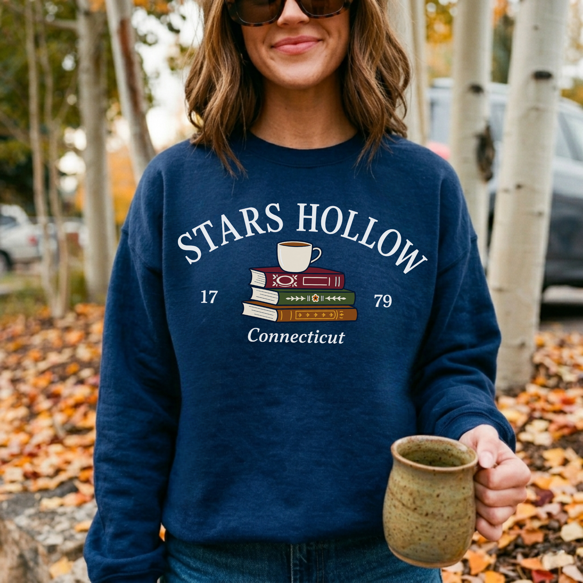 Woman wearing a navy Stars Hollow Connecticut sweatshirt holding a coffee mug among colorful fall leaves.
