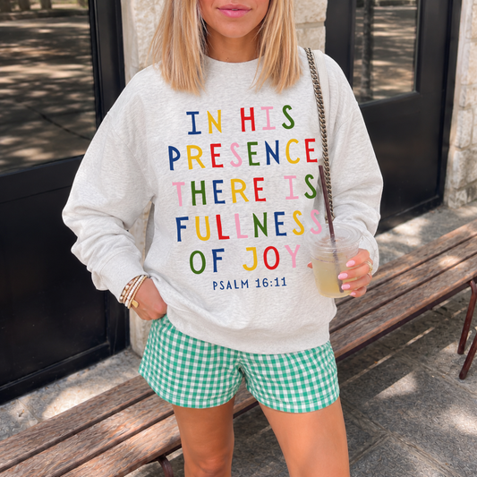 Woman wearing an oatmeal heather Christian crewneck sweatshirt with colorful Psalm 16:11 Scripture design while holding a drink outdoors.