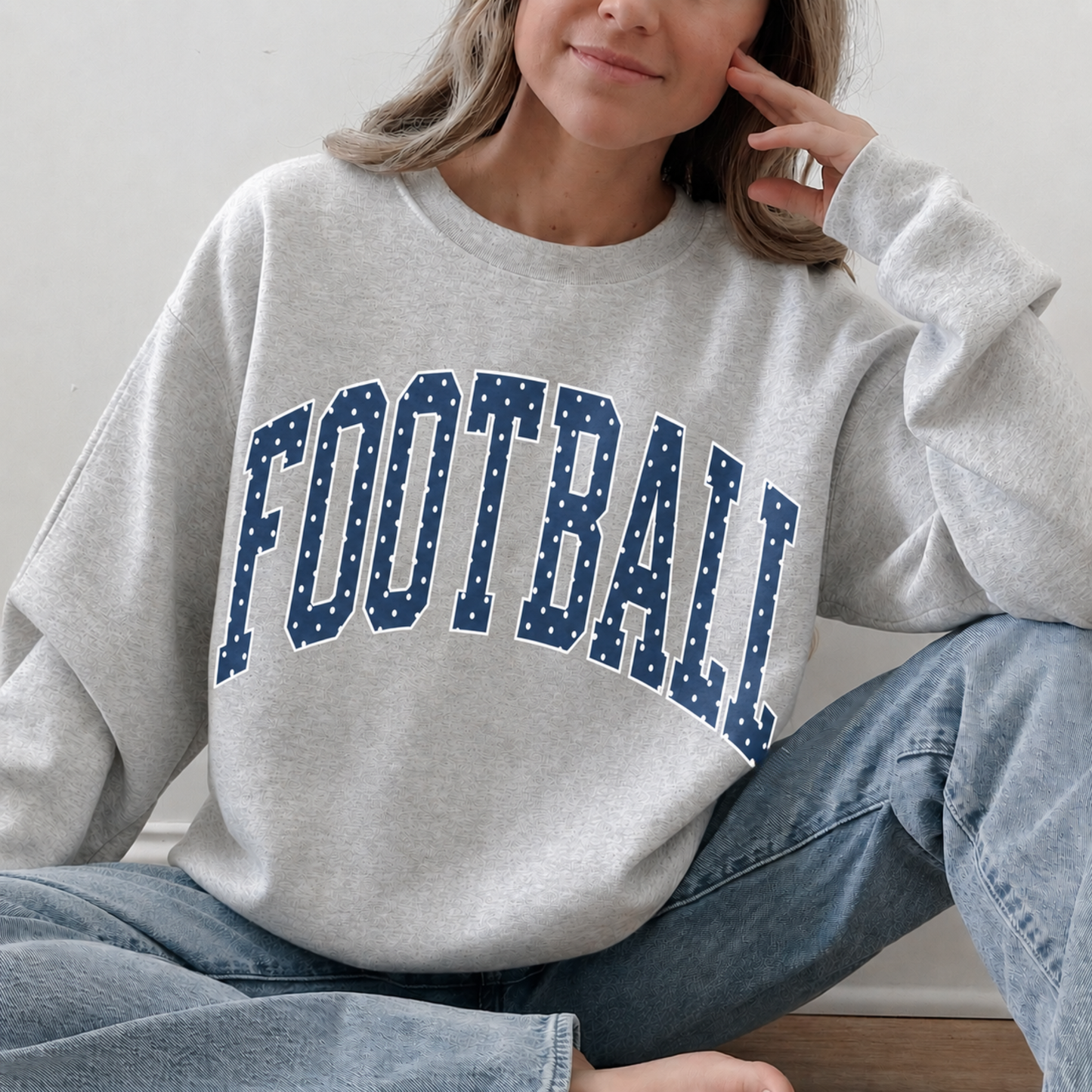 Woman wearing heather grey football sweatshirt with navy polka dot varsity lettering and jeans