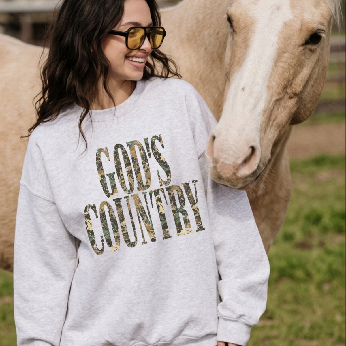 Woman wearing a heather grey God’s Country camo crewneck sweatshirt beside a horse in a country setting