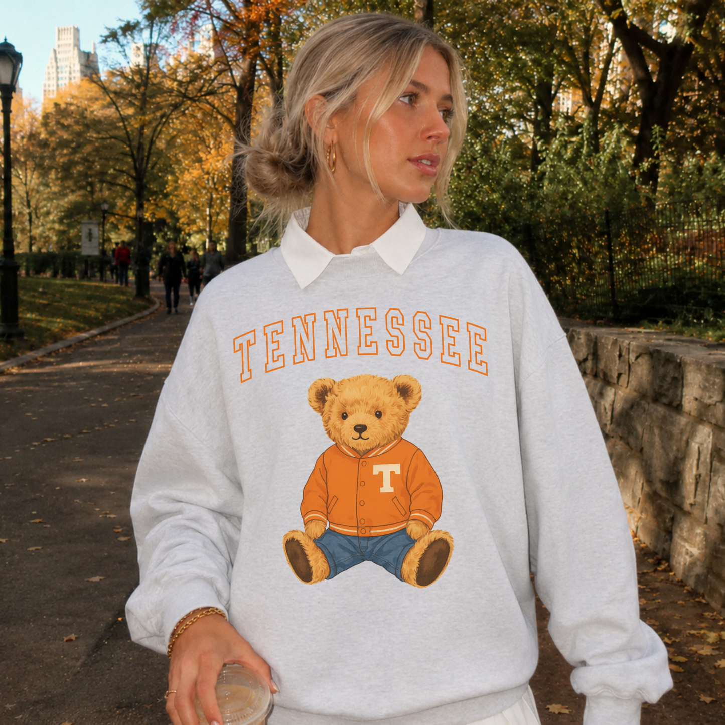 Woman wearing an oatmeal heather Lane Seven crewneck sweatshirt featuring an updated Tennessee varsity arch with a teddy bear wearing an orange varsity jacket during a fall walk.