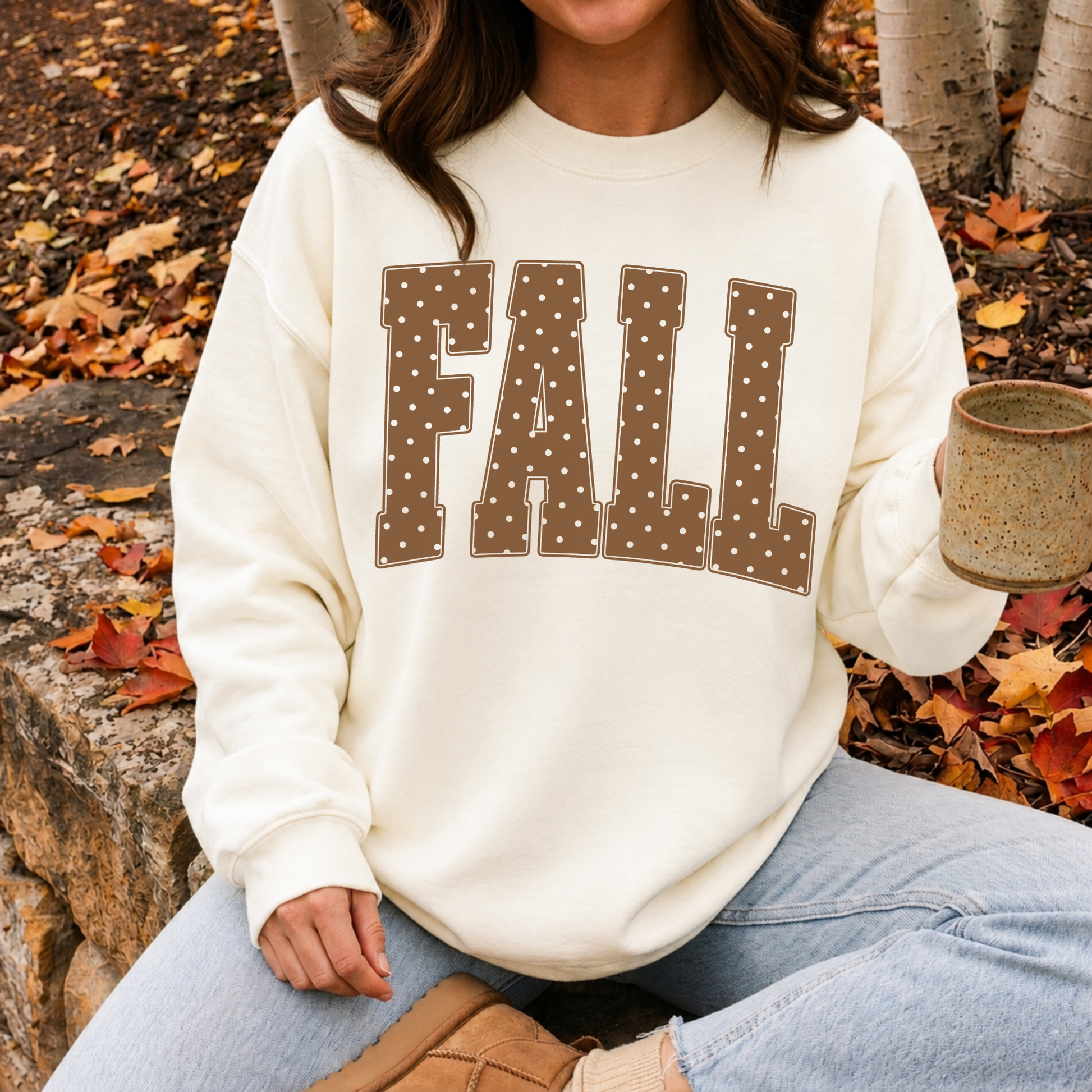 Close-up of a cream fall sweatshirt featuring oversized polka dot "FALL" lettering paired with jeans and a coffee mug for a cozy autumn aesthetic.