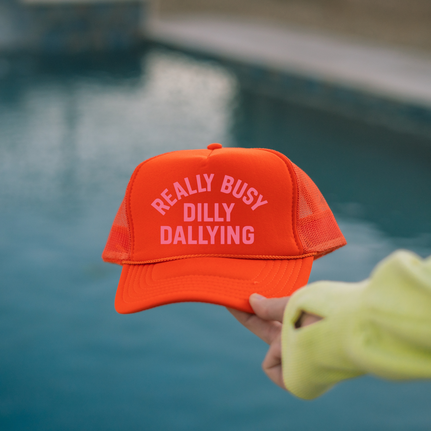 Orange Really Busy Dilly Dallying trucker hat held near pool water