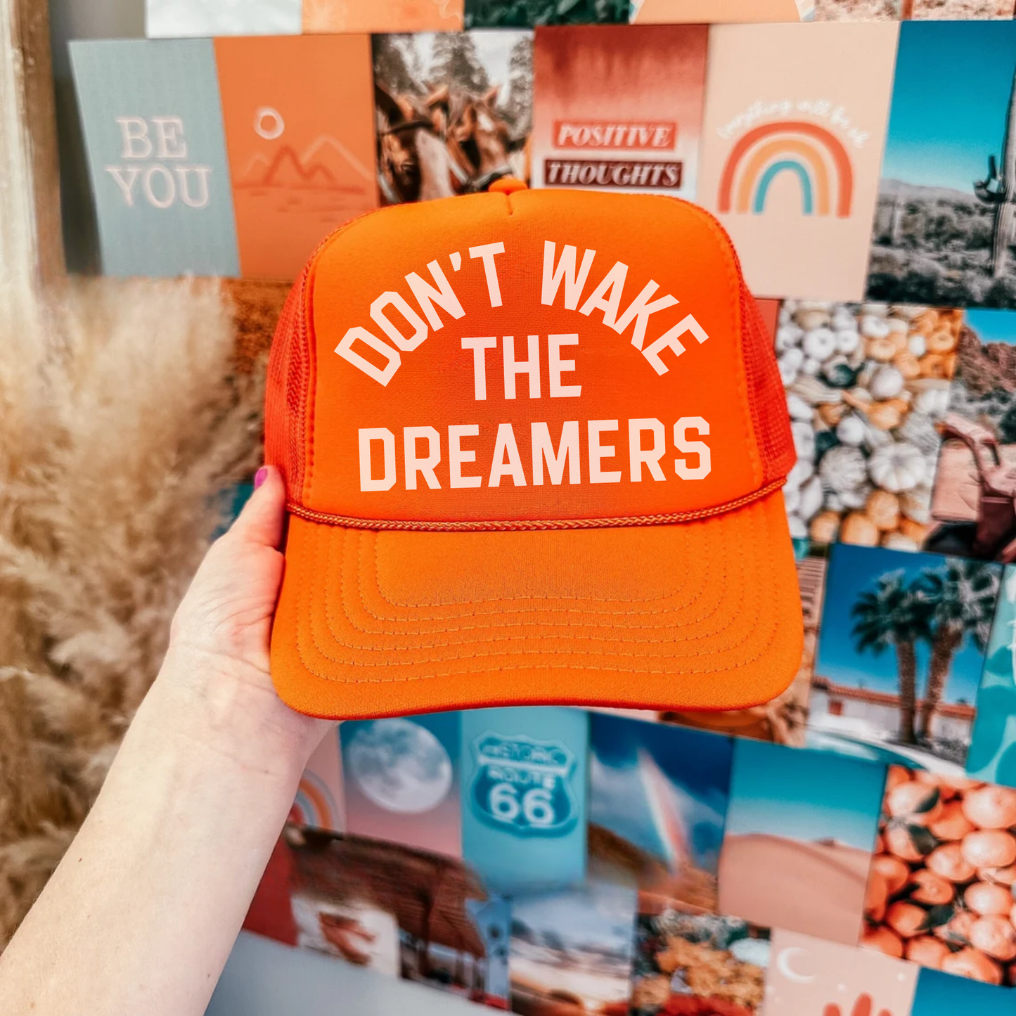 An orange trucker hat with the text 'DON'T WAKE THE DREAMERS' printed on the front, held in a hand against a background with various colorful images and objects.