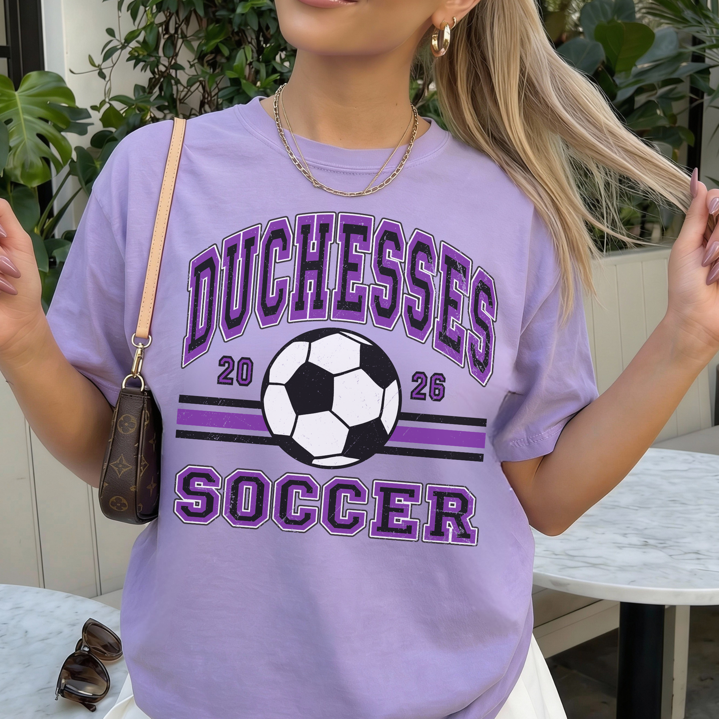 Woman wearing purple Duchesses Soccer 2026 shirt styled with purse and jewelry