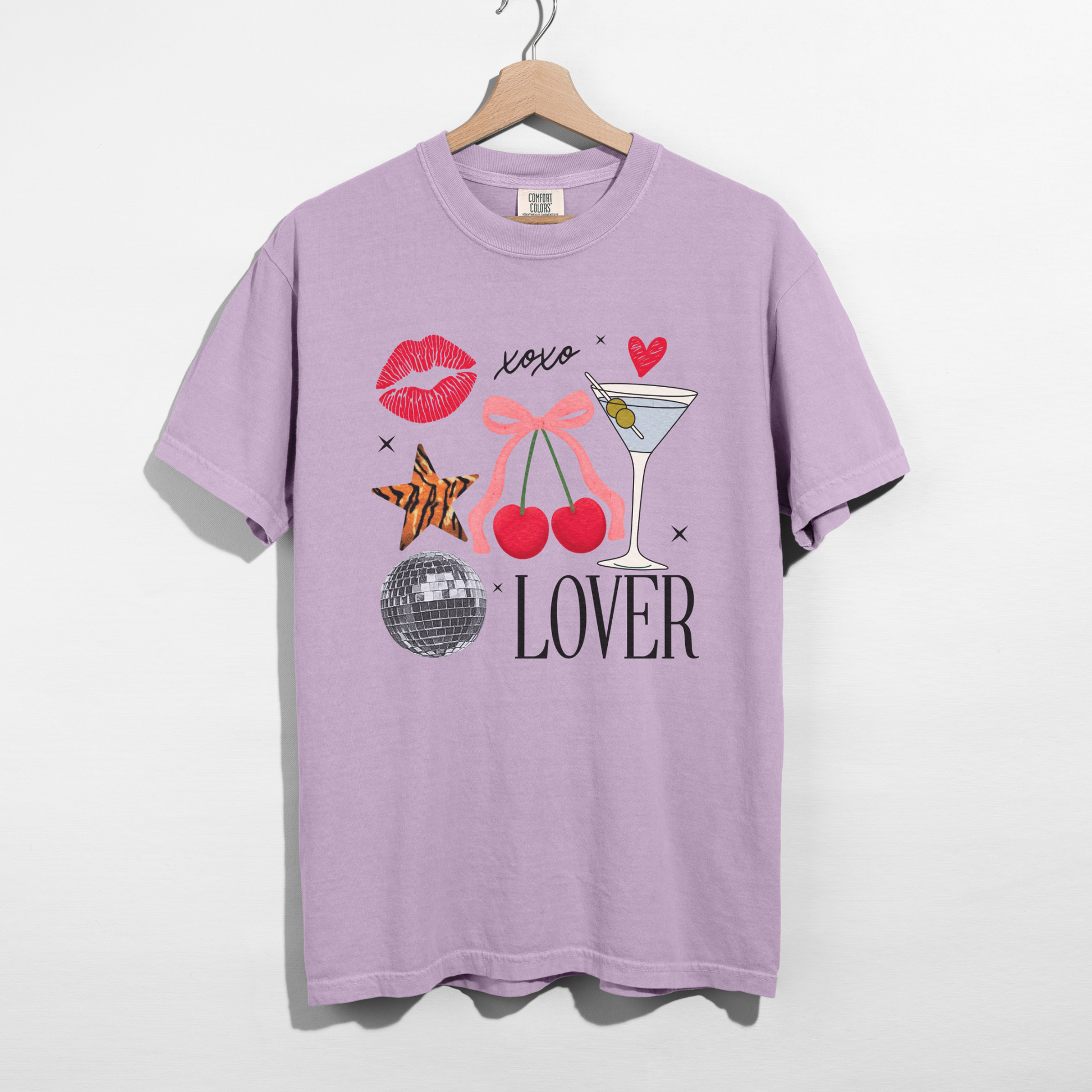 Lavender Comfort Colors Lover graphic tee with cherries, bow, martini glass, and disco ball design hanging on wooden hanger.