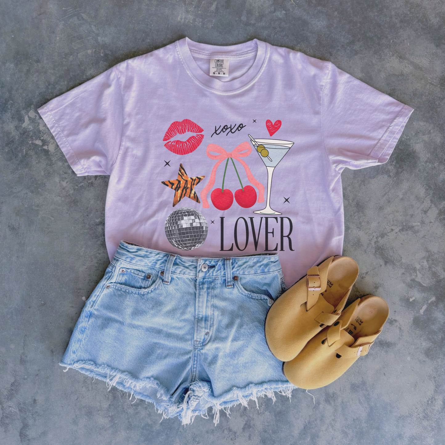 Flat lay of lavender Lover graphic tee paired with denim shorts and tan Birkenstock-style sandals.