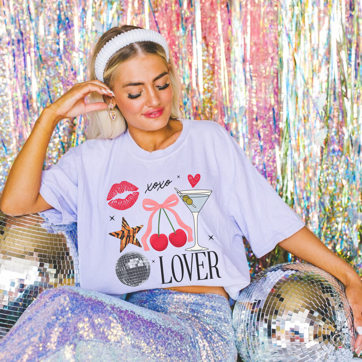 Blonde woman wearing lavender Lover graphic tee surrounded by disco balls and pink tinsel backdrop, fun retro vibe.