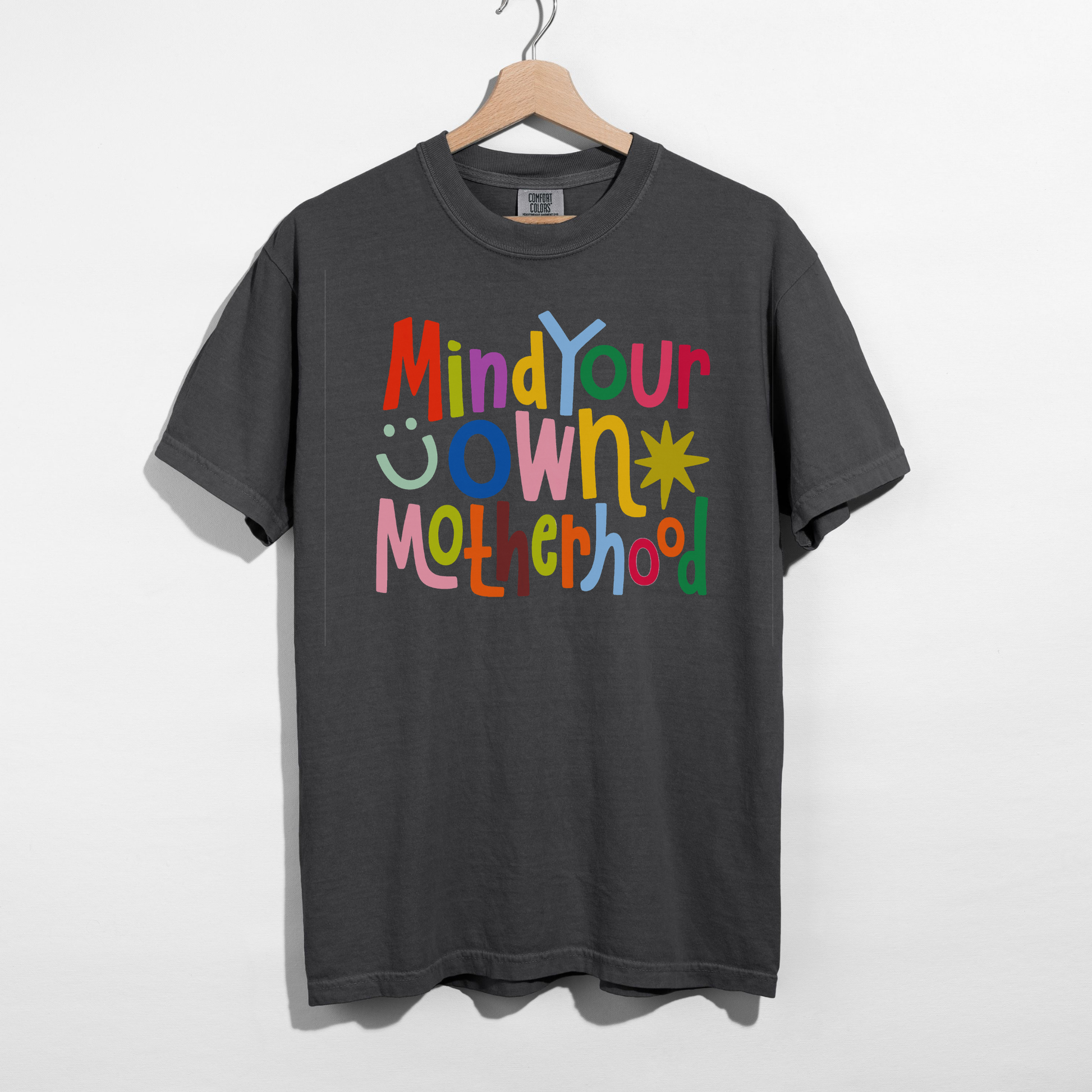 Colorful Mind Your Own Motherhood graphic printed on a pepper Comfort Colors unisex t-shirt hanging on wooden hanger