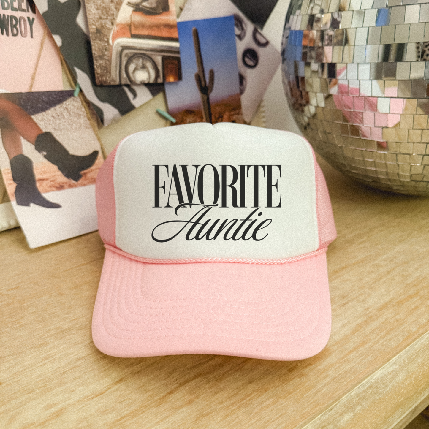 Pink and white cap with 'Favorite Auntie' text on a wooden surface with photos and a disco ball.