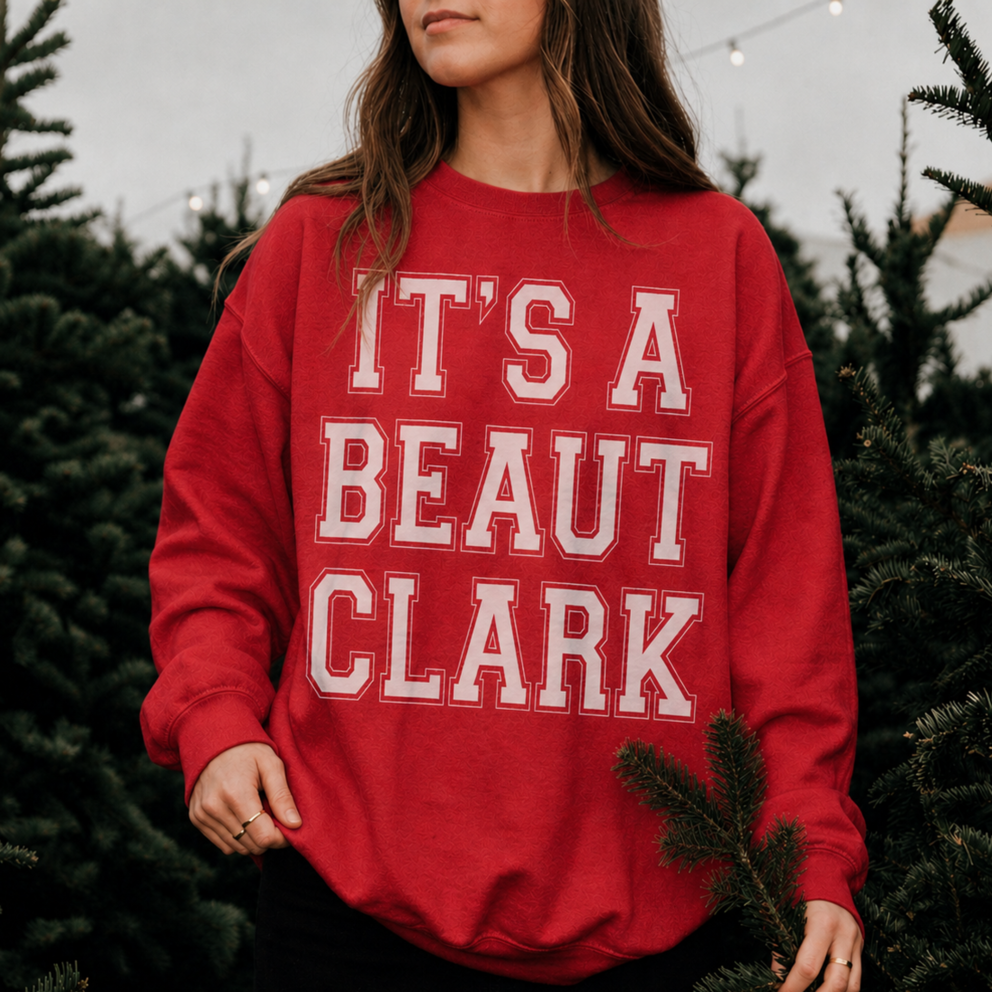 Woman wearing red It's a Beaut Clark Christmas sweatshirt with white graphic at a festive Christmas tree farm.