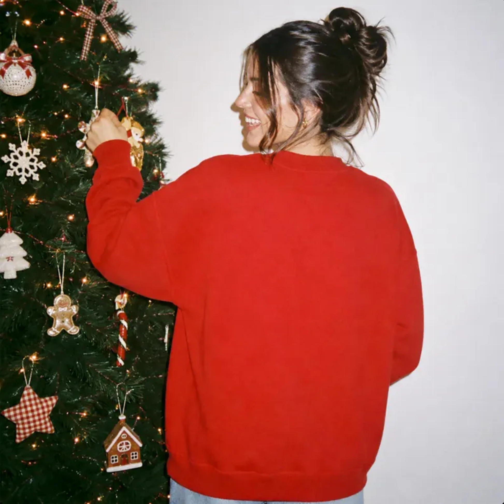 Back view of woman wearing an oversized red Christmas crewneck sweatshirt while decorating a Christmas tree.