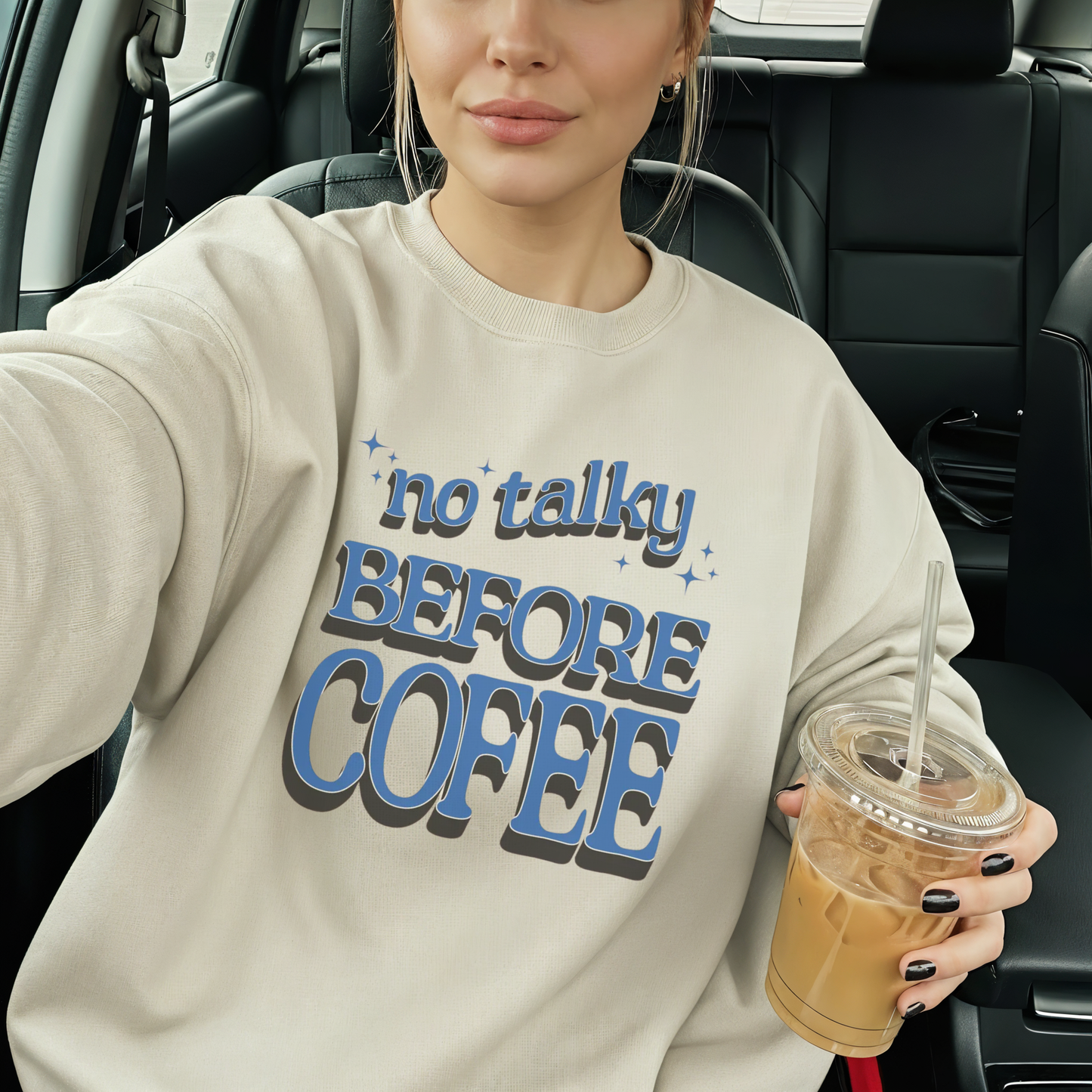 Woman wearing sand ‘No Talky Before Coffee’ sweatshirt holding iced coffee in car