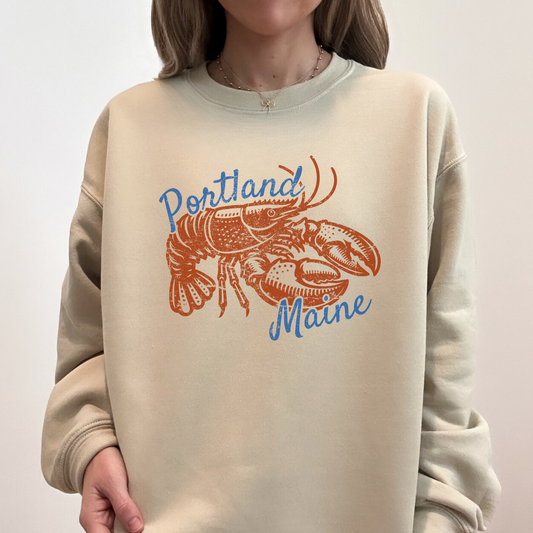 Woman wearing sand-colored Portland Maine sweatshirt with vintage lobster print and blue lettering.