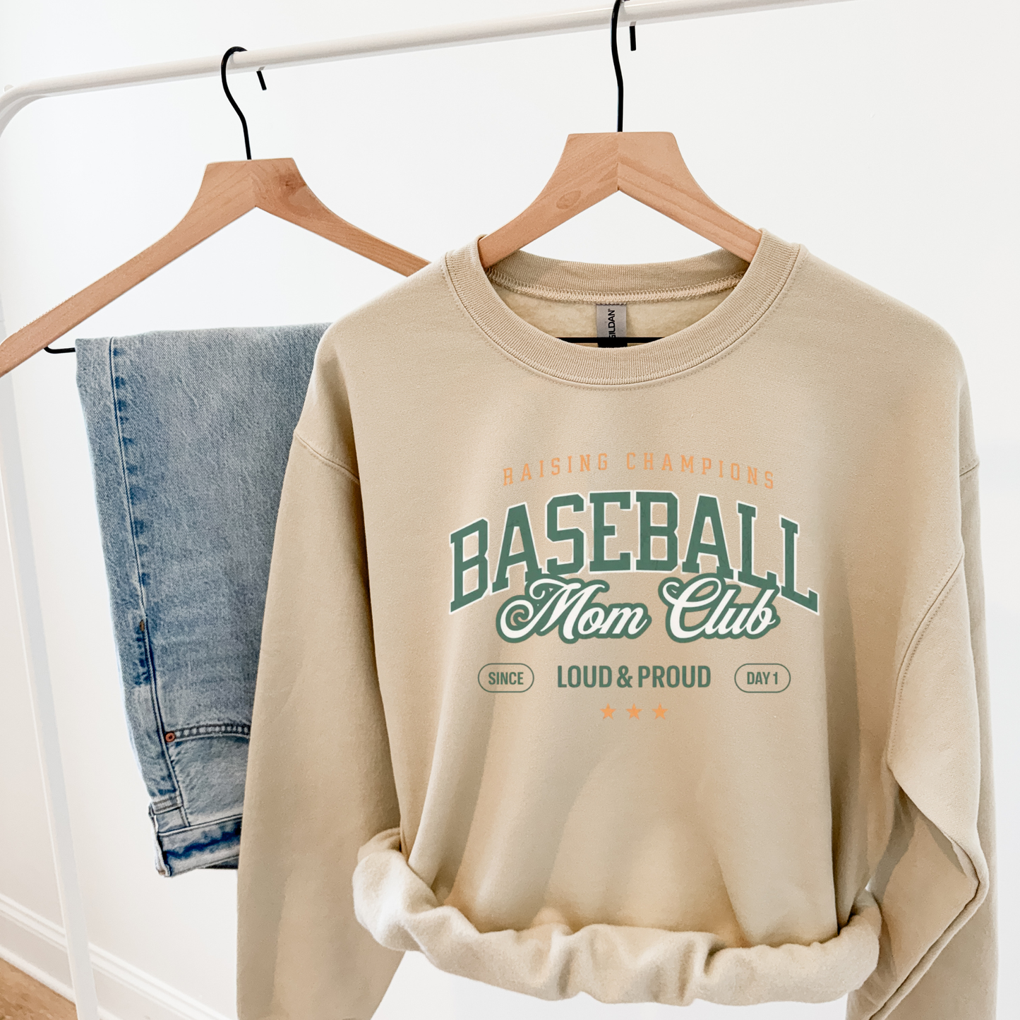 Sand “Baseball Mom Club” sweatshirt hanging on a clothing rack next to denim jeans.