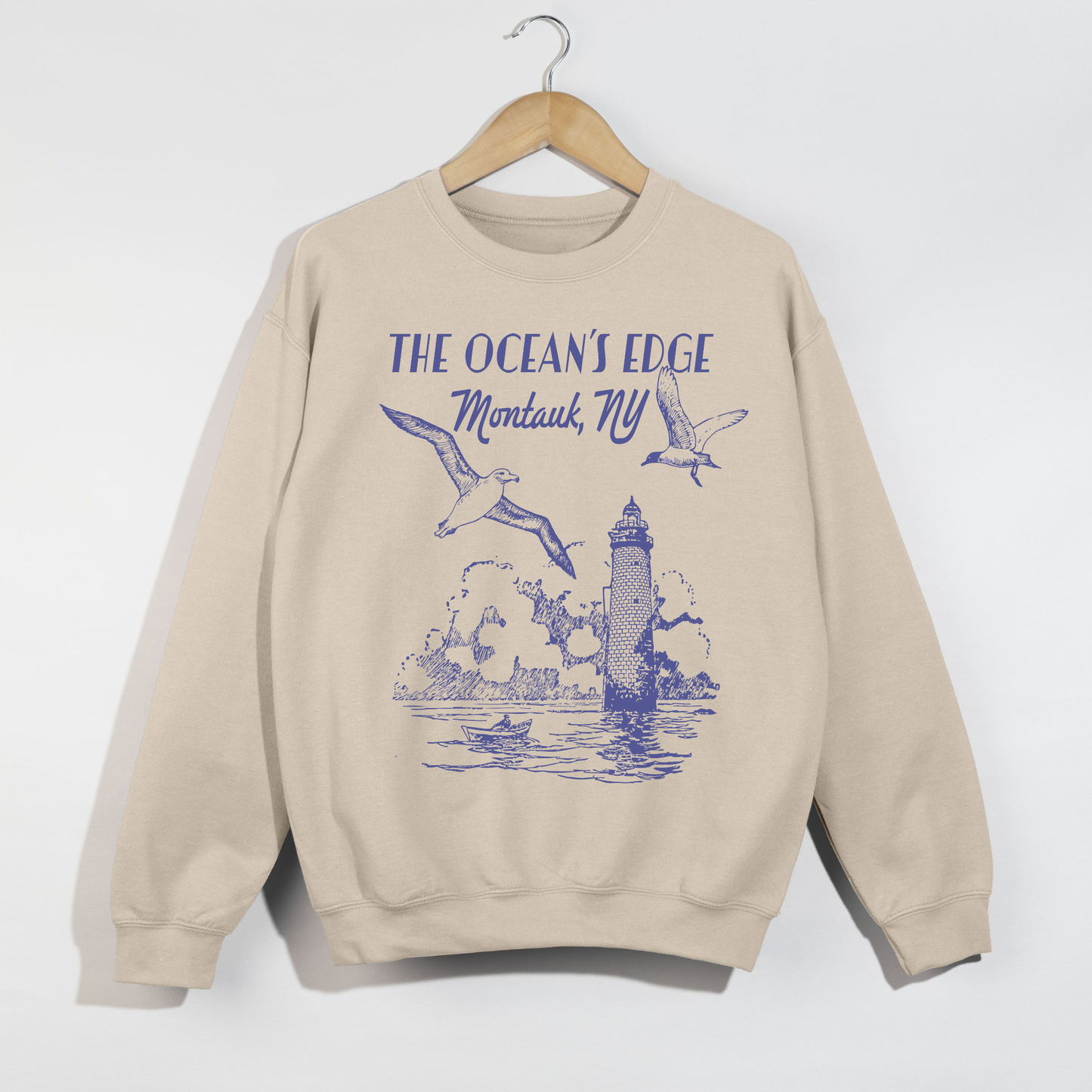 "Sand-colored Montauk coastal lighthouse graphic sweatshirt hanging on wooden hanger."