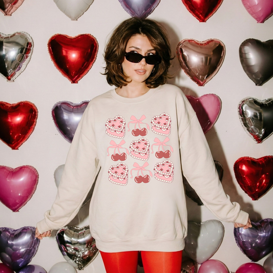 Woman wearing a sand-colored Valentine sweatshirt with pink heart cake and cherry graphic design, posing in front of metallic heart balloons.
