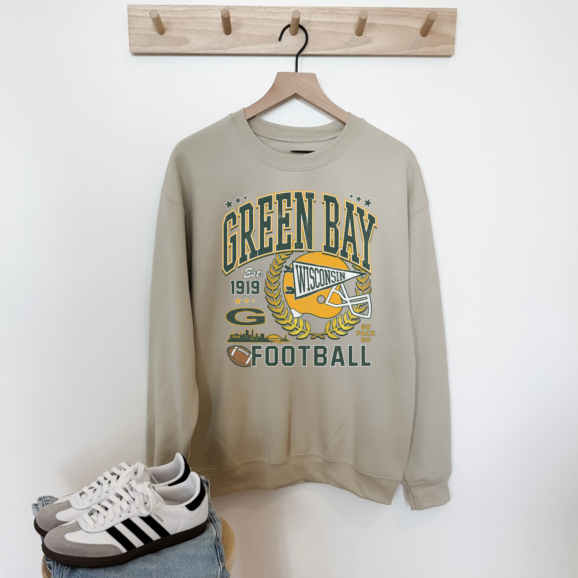 Beige sweatshirt with 'Green Bay Football' design on a hanger, with sneakers and jeans at the bottom.