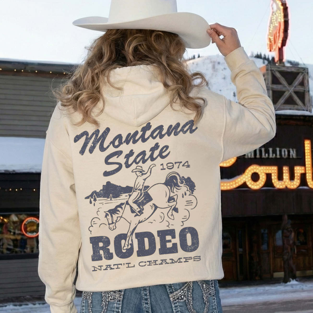 Woman wearing sand Montana State Rodeo hoodie with oversized vintage western cowboy back graphic and white cowboy hat.