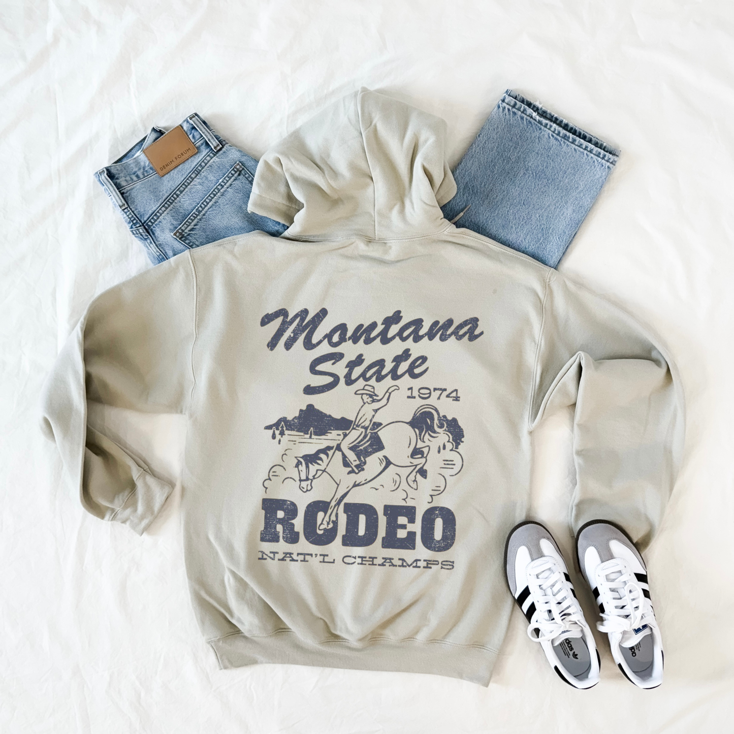 Beige hoodie with 'Montana State Rodeo Nat'l Champs' design, blue jeans, and white sneakers on a white background