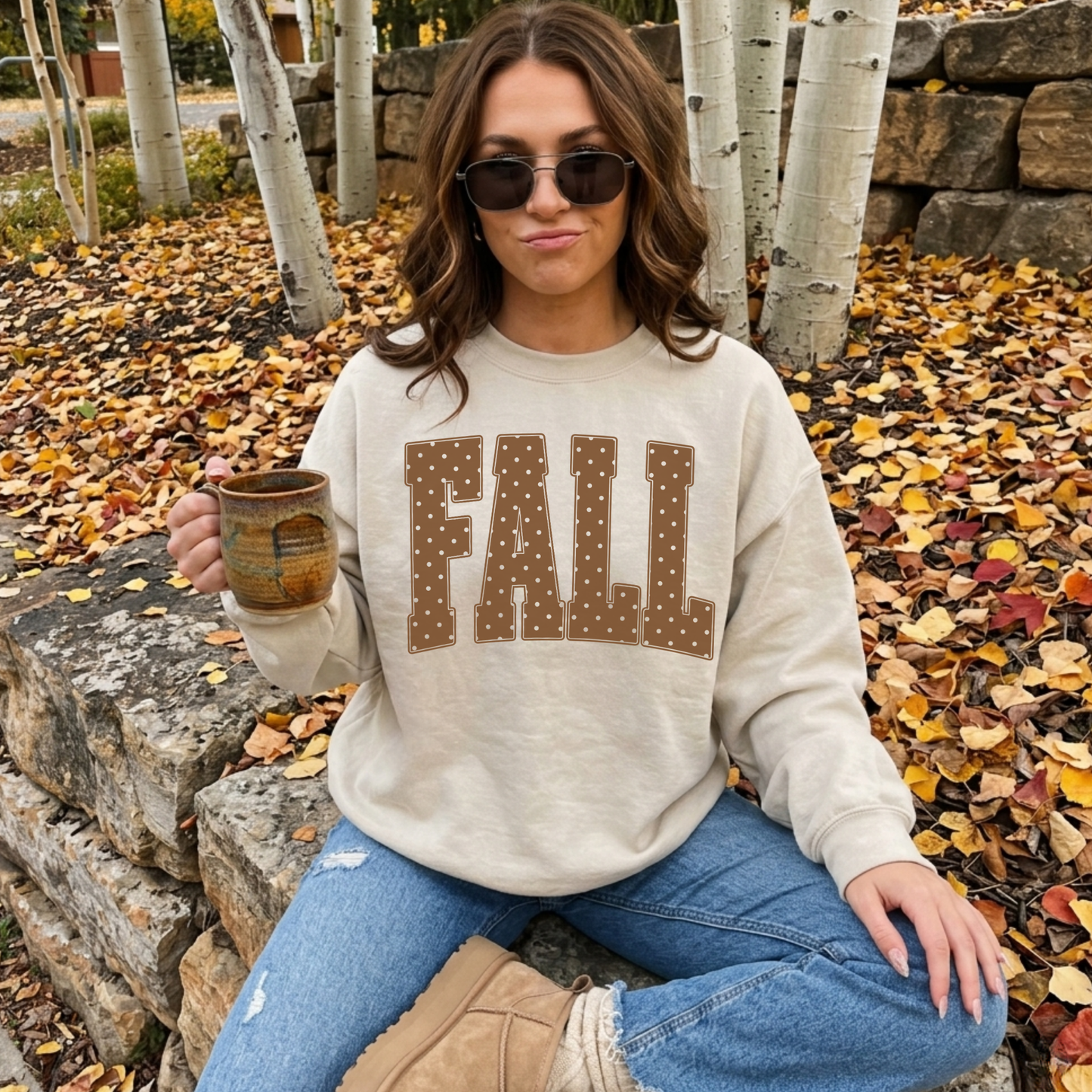 Woman wearing a neutral cream fall graphic crewneck sweatshirt with oversized polka dot "FALL" lettering while enjoying coffee outdoors among colorful autumn leaves.