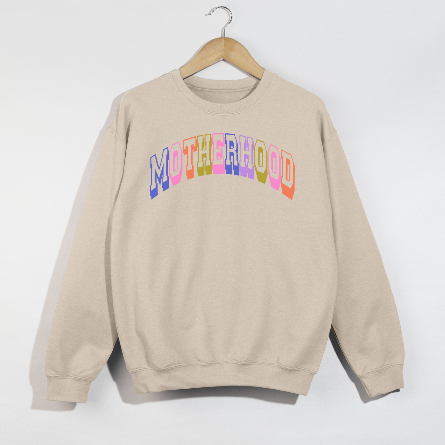 Sand retro motherhood sweatshirt with colorful collegiate lettering hanging on wooden hanger against white wall.