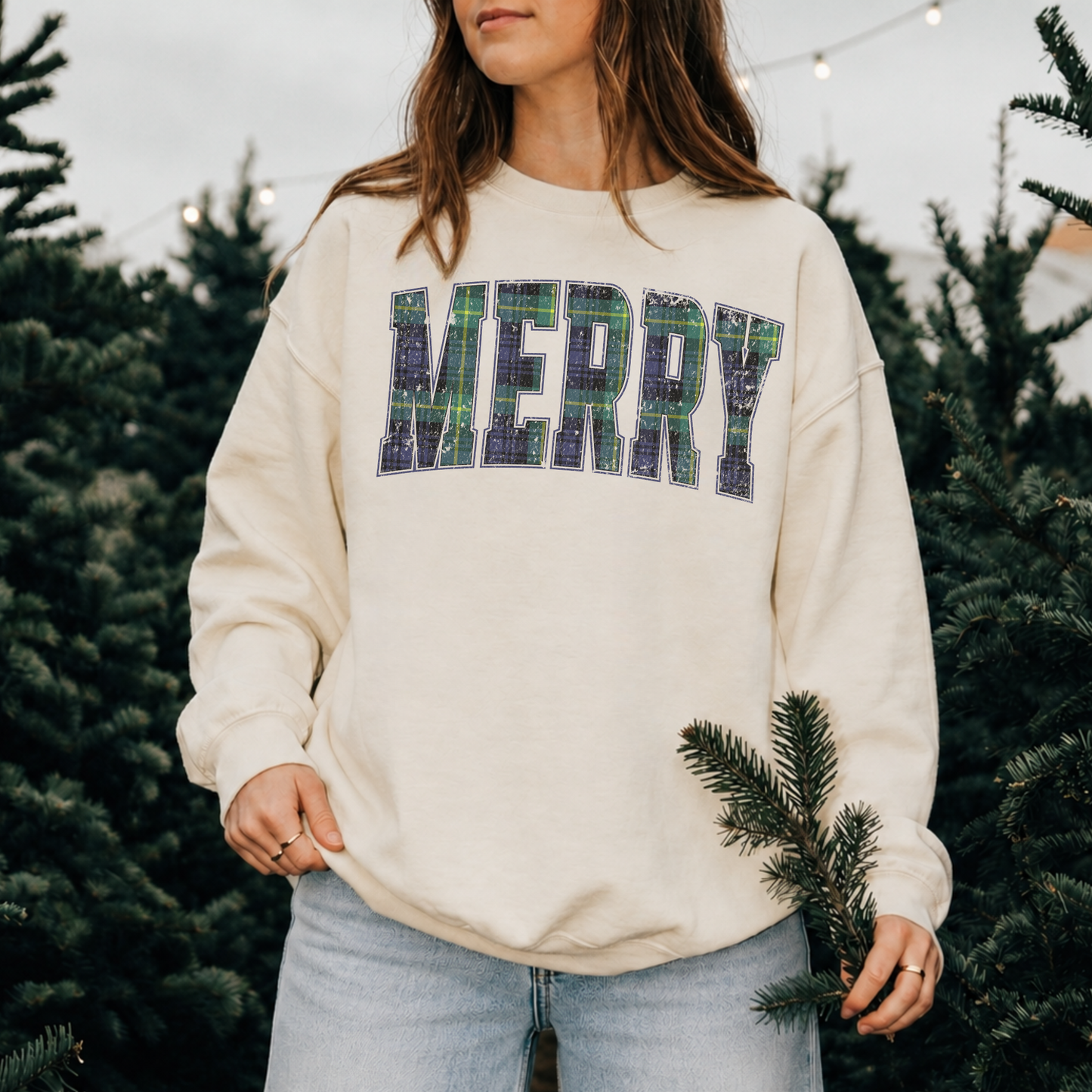 Woman wearing a sandshell Lane Seven crewneck sweatshirt featuring distressed navy and green plaid "MERRY" lettering while walking through a Christmas tree farm.