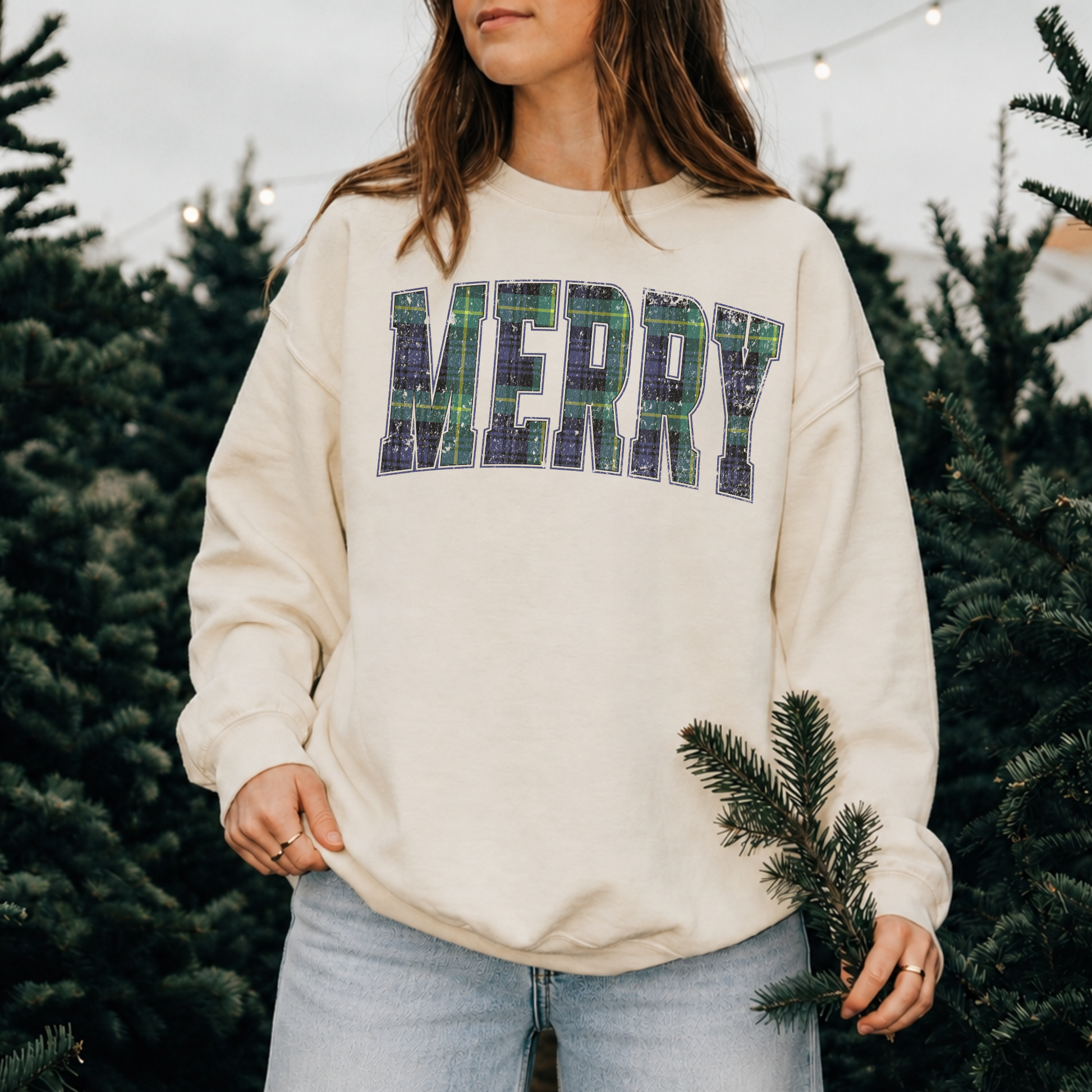 Woman wearing a sandshell Lane Seven crewneck sweatshirt featuring distressed navy and green plaid "MERRY" lettering while walking through a Christmas tree farm.
