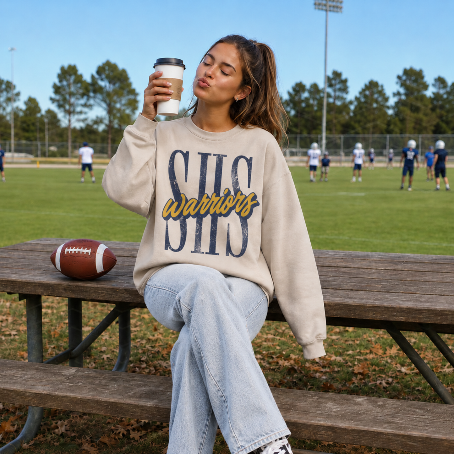 Woman wearing a Sterling Warriors SHS graphic sweatshirt at a football field featuring oversized distressed SHS lettering with navy and gold Warriors script.