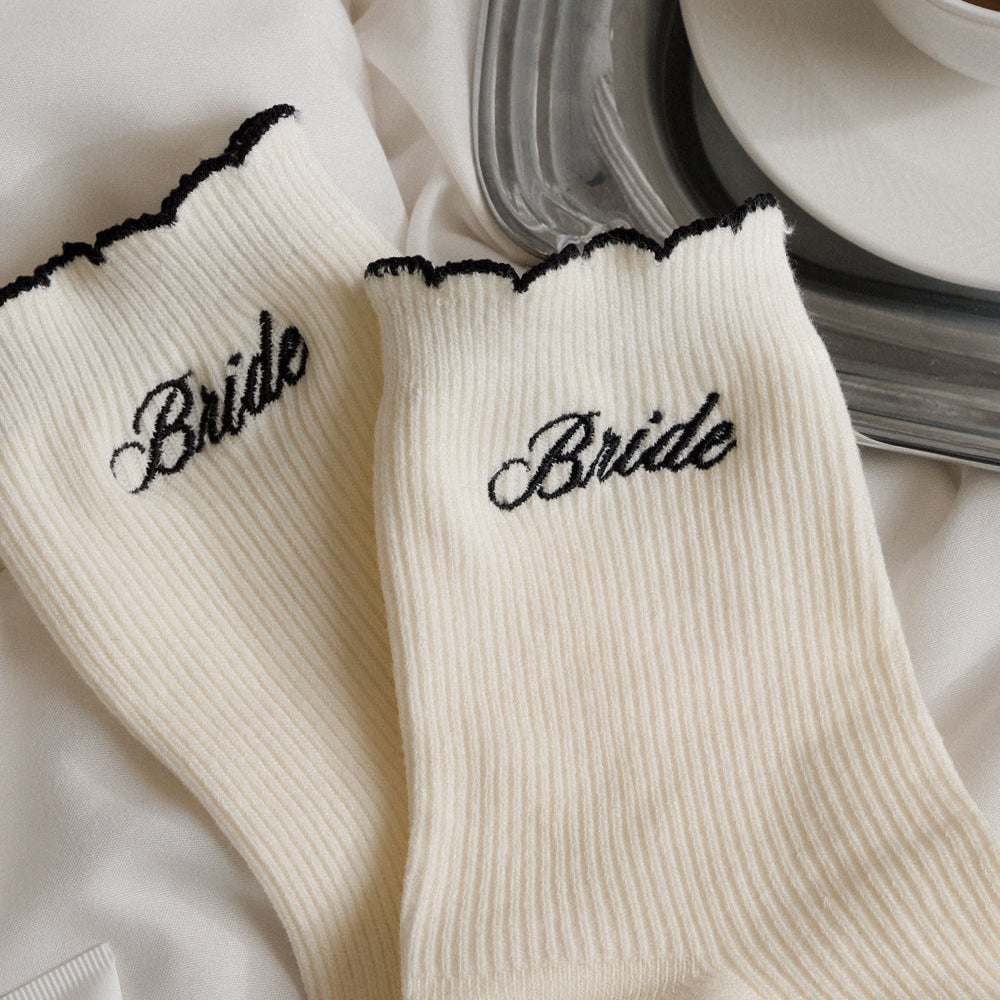 White socks with 'Bride' embroidery on a white surface with a cup of coffee in the background.