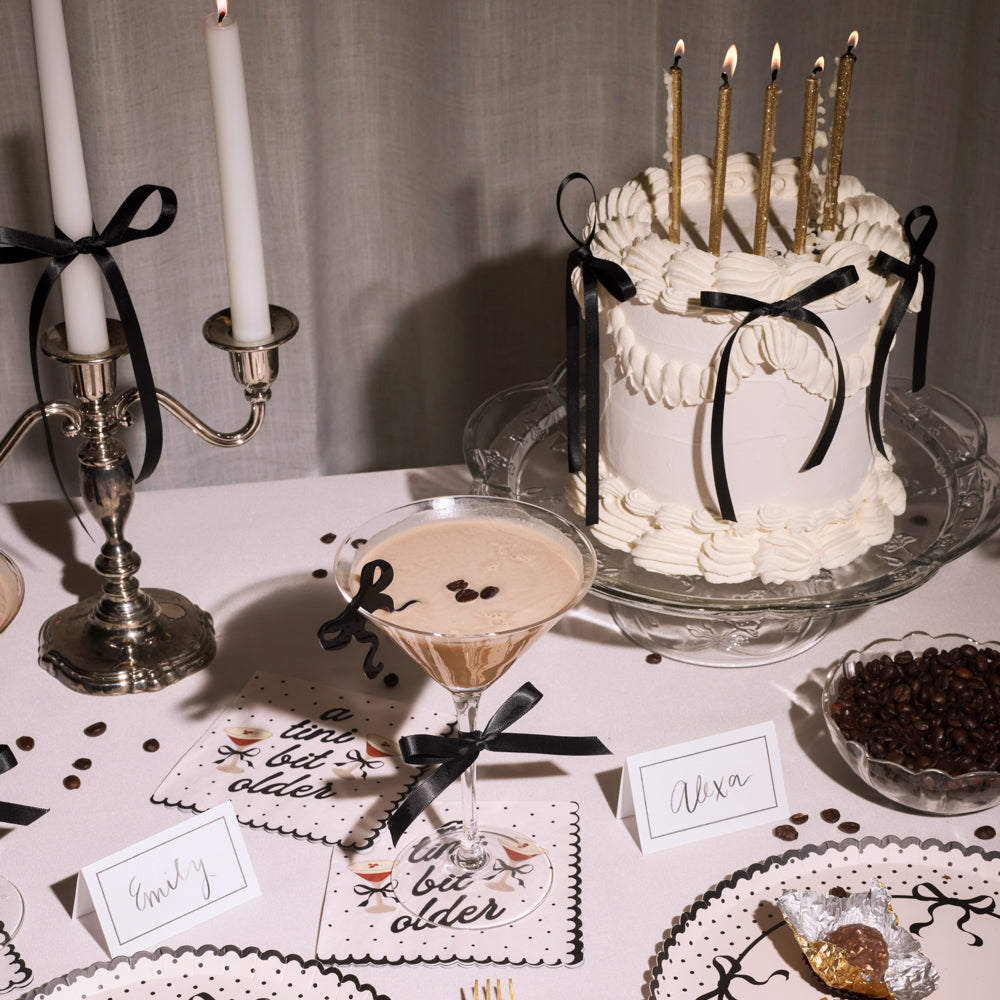 Decorative table setting with a cake, candles, and place cards, and espresso martini napkins on a white tablecloth.