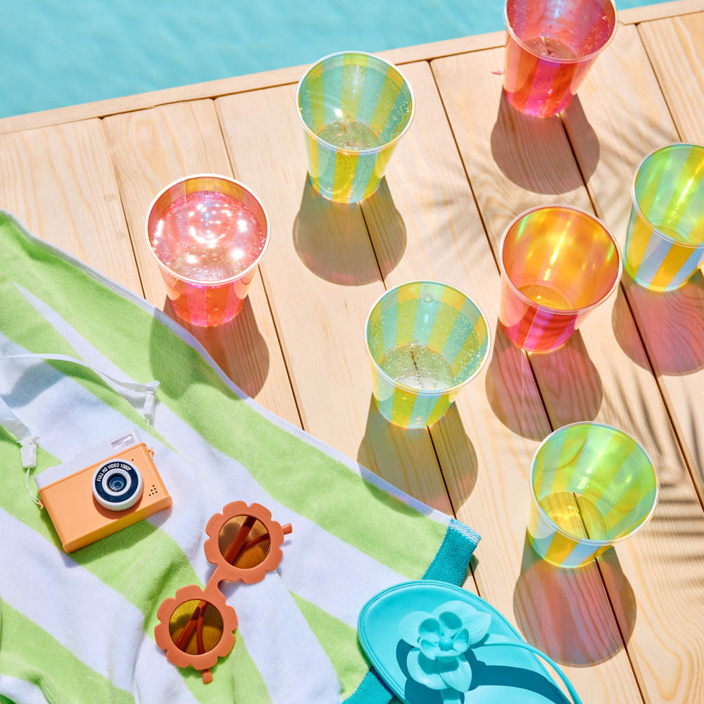 Colorful plastic cups on a wooden deck near a pool, with a camera, sunglasses, and flip-flops.