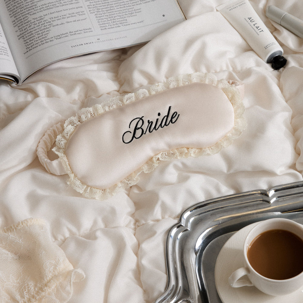 Sleep mask with 'Bride' printed on it, next to an open book and a cup of coffee on a tray.