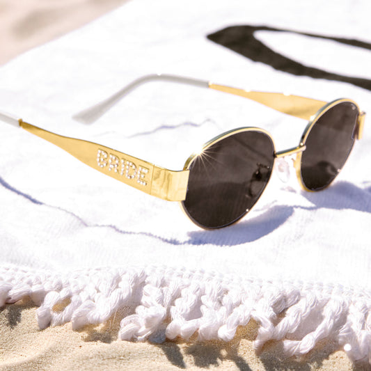 Gold-framed sunglasses with dark lenses on a white towel at the beach