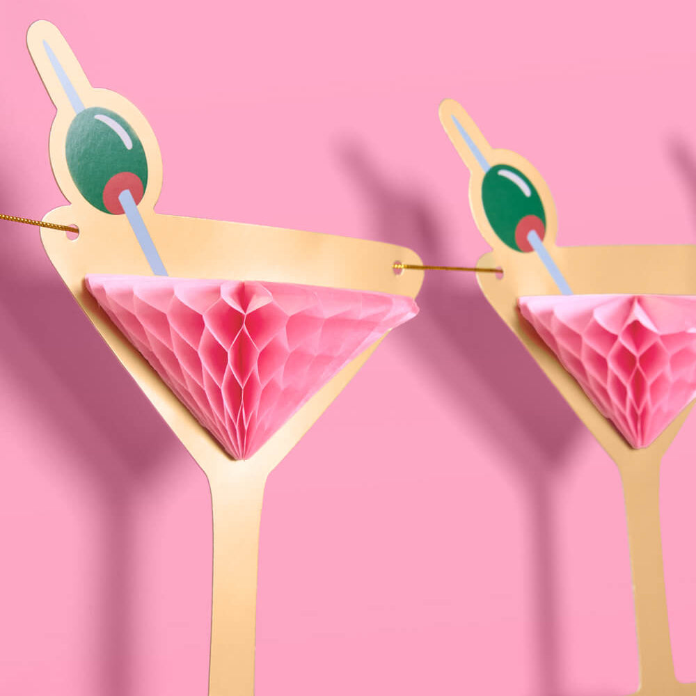 Two paper cocktail decorations with pink drink and green olive on a pink background