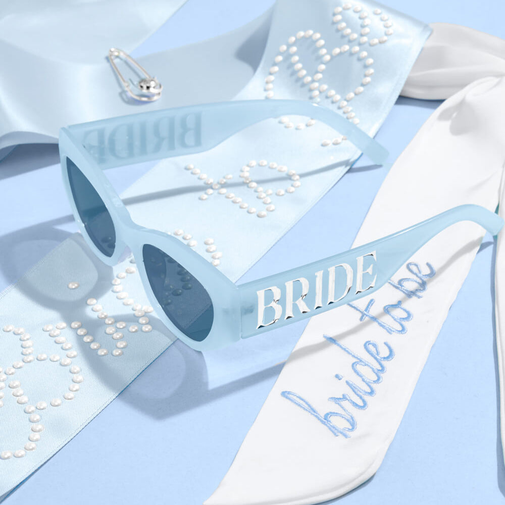 Clear sunglasses with 'Bride' text and a white sandal with 'bride to be' text on a light blue background.