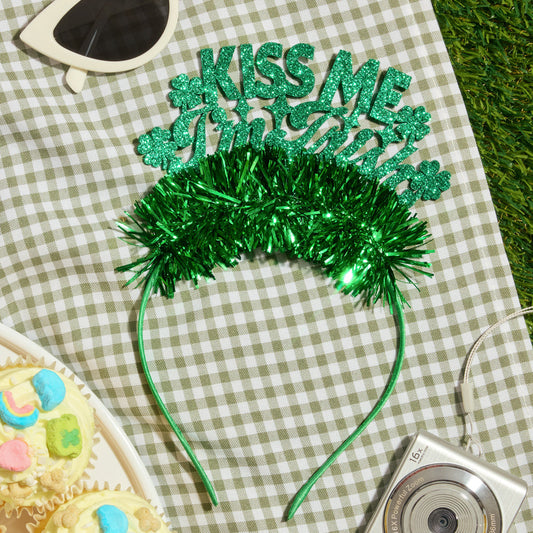 Green glittery 'Kiss Me' headband on a checkered tablecloth with sunglasses, camera, and cupcakes.