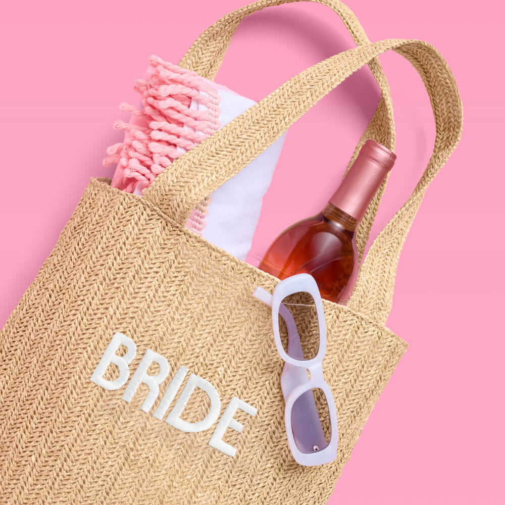 Woven bag labeled 'BRIDE' with a bottle, sunglasses, and hairbrush on a pink background