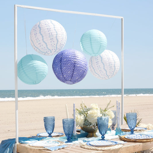 Outdoor dining setup with paper lanterns, blue glasses, and white flowers by the beach.