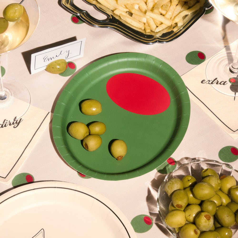 Green paper plate with red dot and olives on a table setting with wine glasses and food.