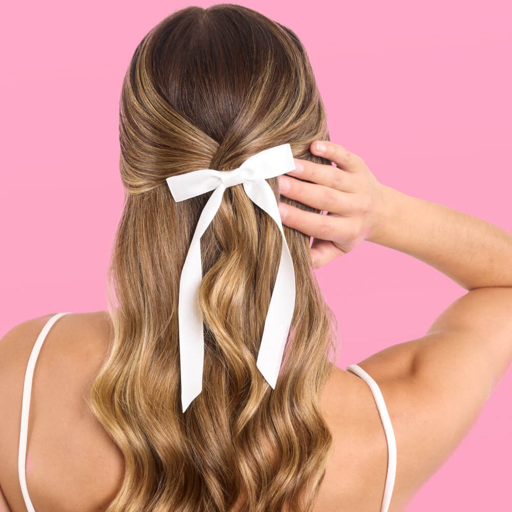 Woman with long, wavy hair tied with a white ribbon against a pink background