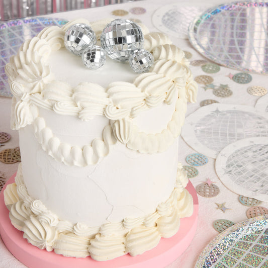 White cake with decorative frosting on a table with disco balls and a patterned tablecloth.