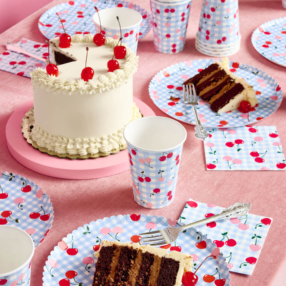 Table setting with cake, cups, and plates featuring cherry design on a pink tablecloth.
