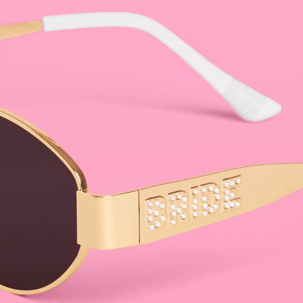 Gold sunglasses with 'Bride' on the arm against a pink background