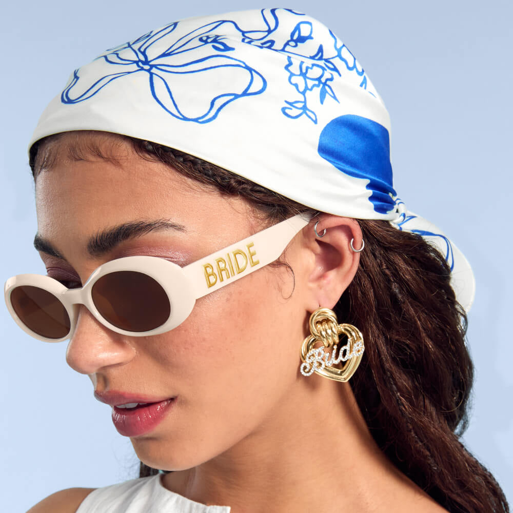 Woman wearing a headscarf with blue patterns, sunglasses with 'Bride' text, and 'girl' earrings against a light blue background.