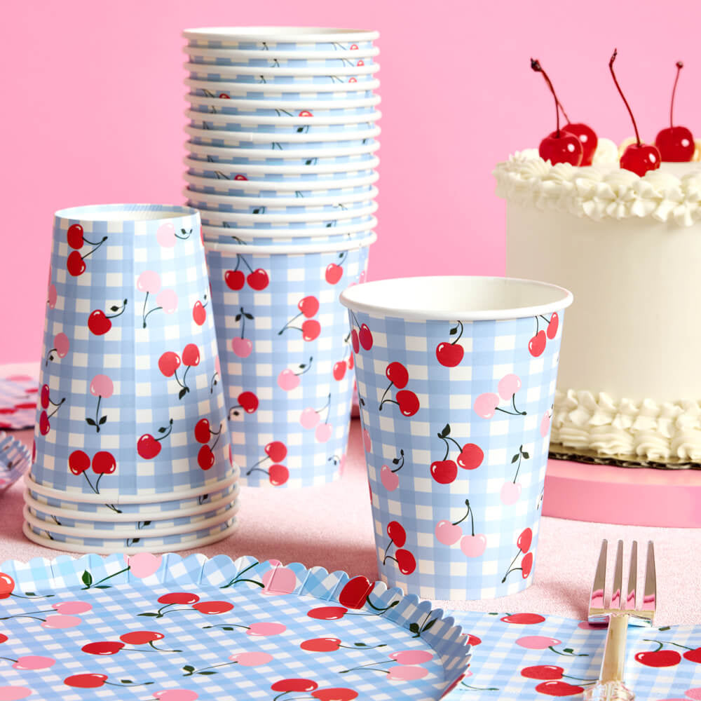 Party supplies with cherry and checkered design on cups, plates, and cake against a pink background