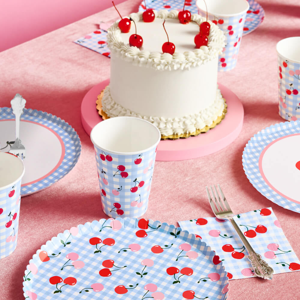 Table setting with cake, cups, and plates featuring cherry design on a pink tablecloth.