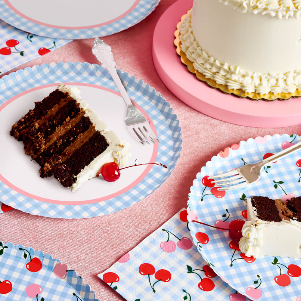 Chocolate cake slices on cherry-themed paper plates and napkins with a pink background.