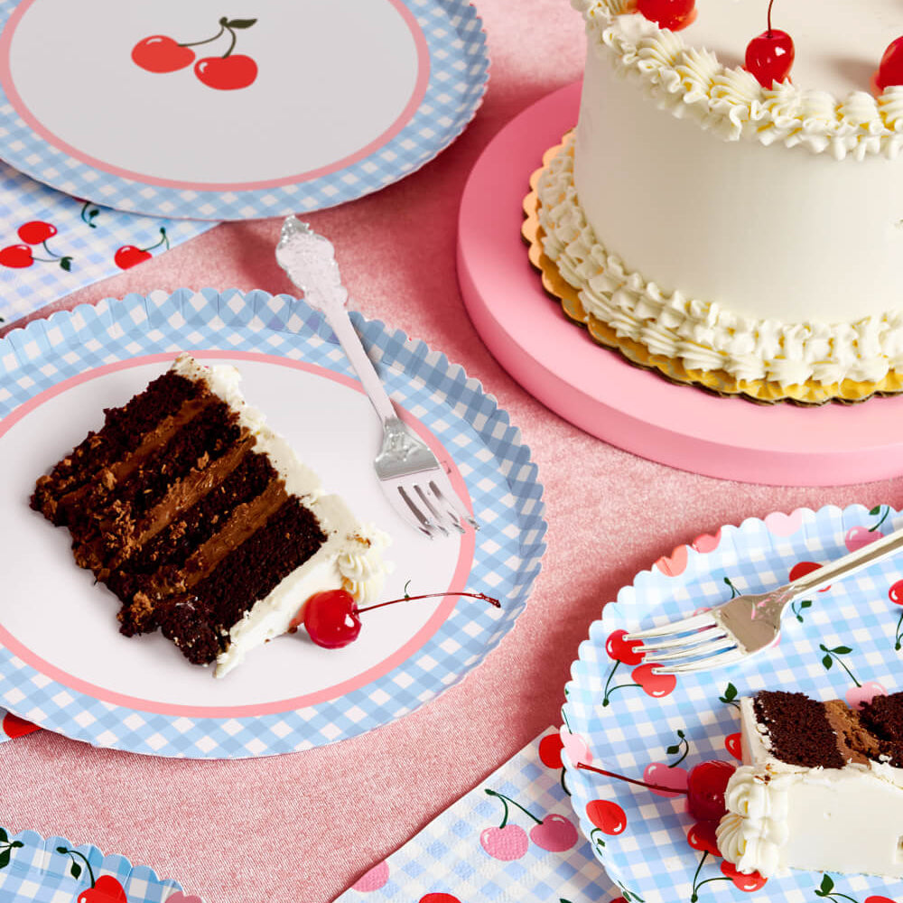 Chocolate cake slices on cherry-themed paper plates and napkins with a pink background.